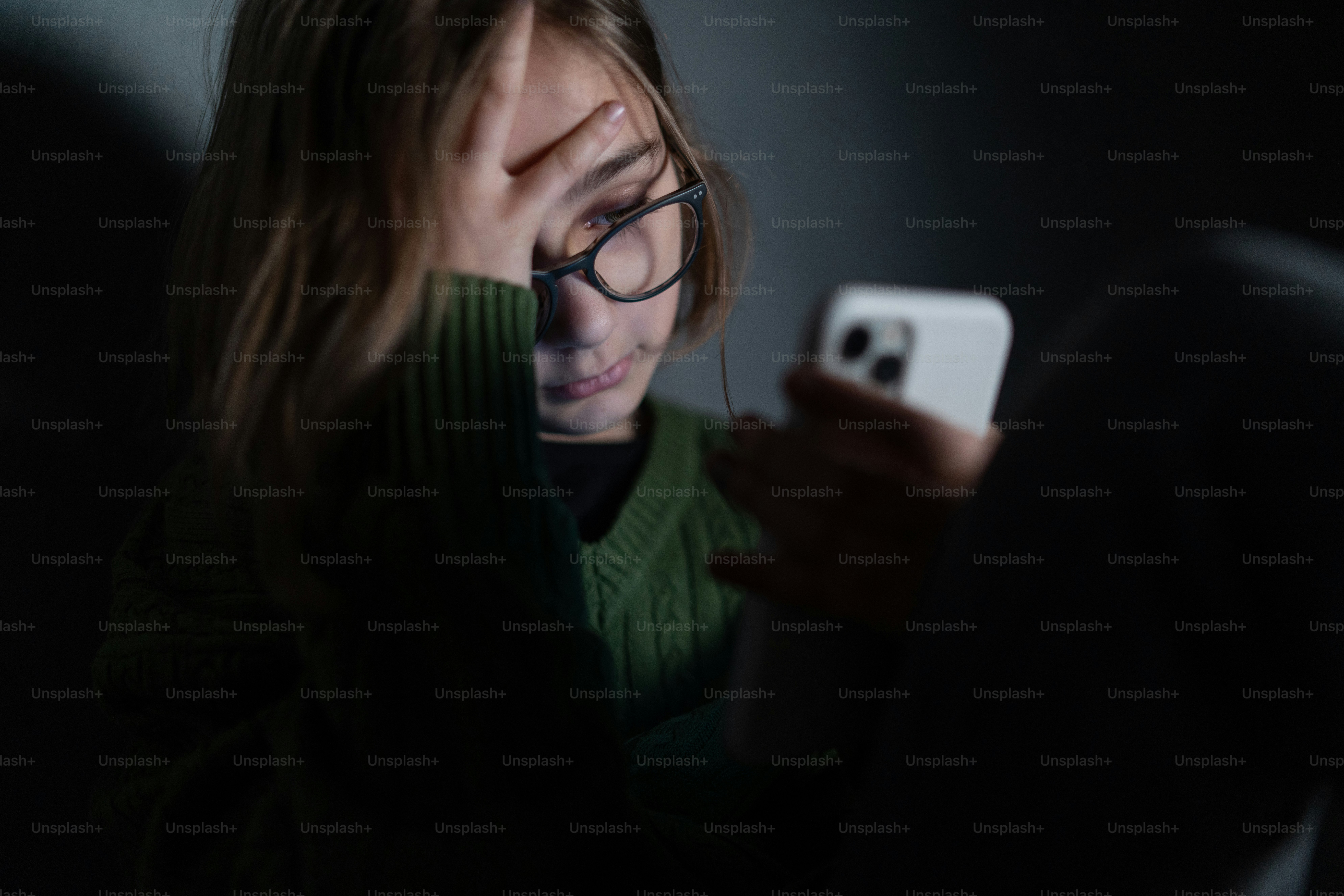A sad little girl, alone in darkness, sitting and using smartphone